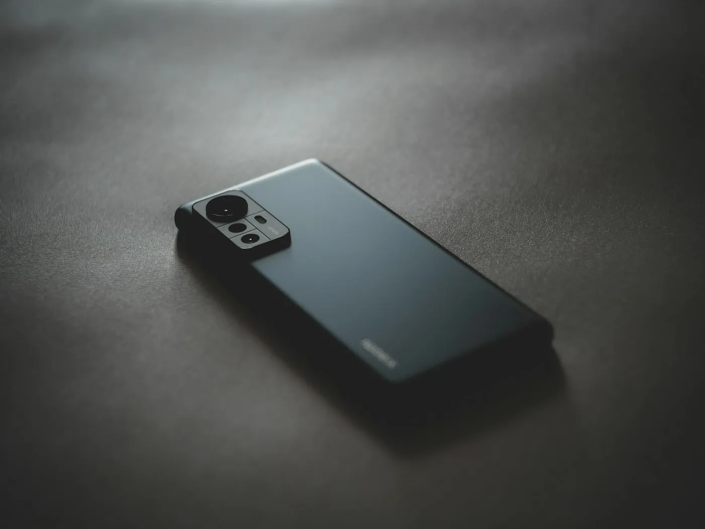 Dark gray smartphone lying face down on a matte surface, its rear cameras in soft light