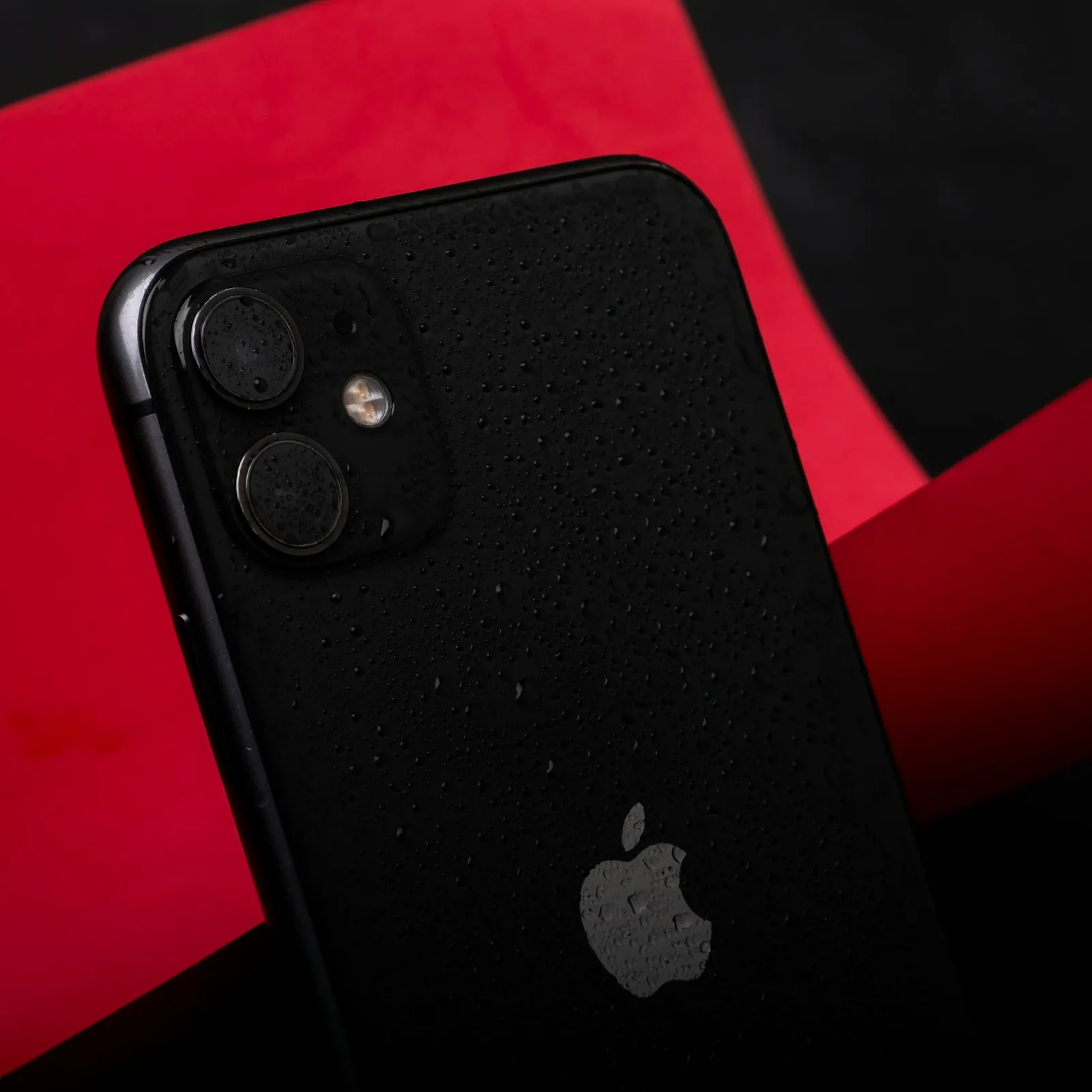 Black smartphone covered in water droplets against folded crimson paper in low-key studio light