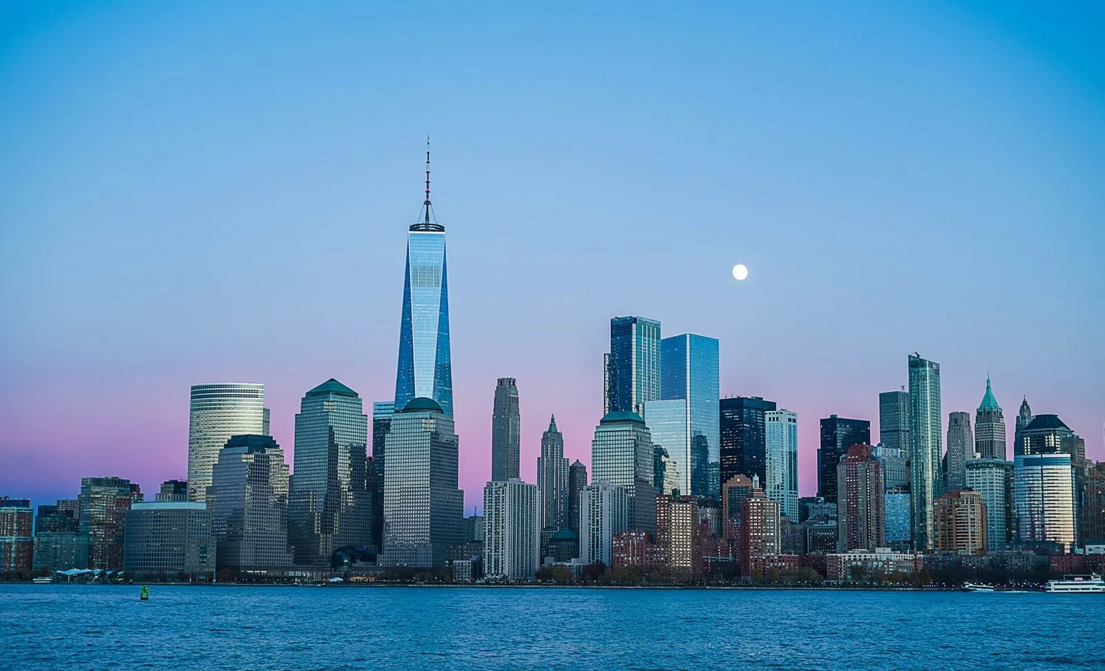 The lit-up skyline of a large financial district at dusk, seen across the water
