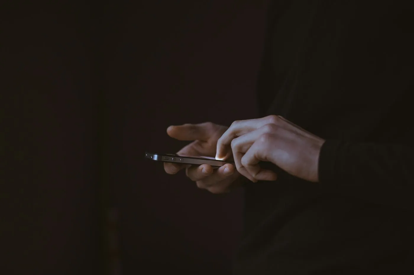 Hands tapping a smartphone screen in a dark room, lit only by the phone's glow