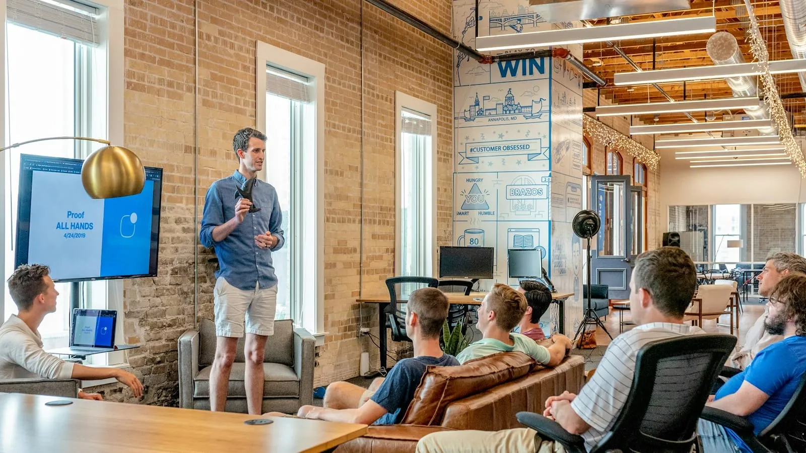 A team gathered around a long office table with laptops while one person presents at the far end of the room