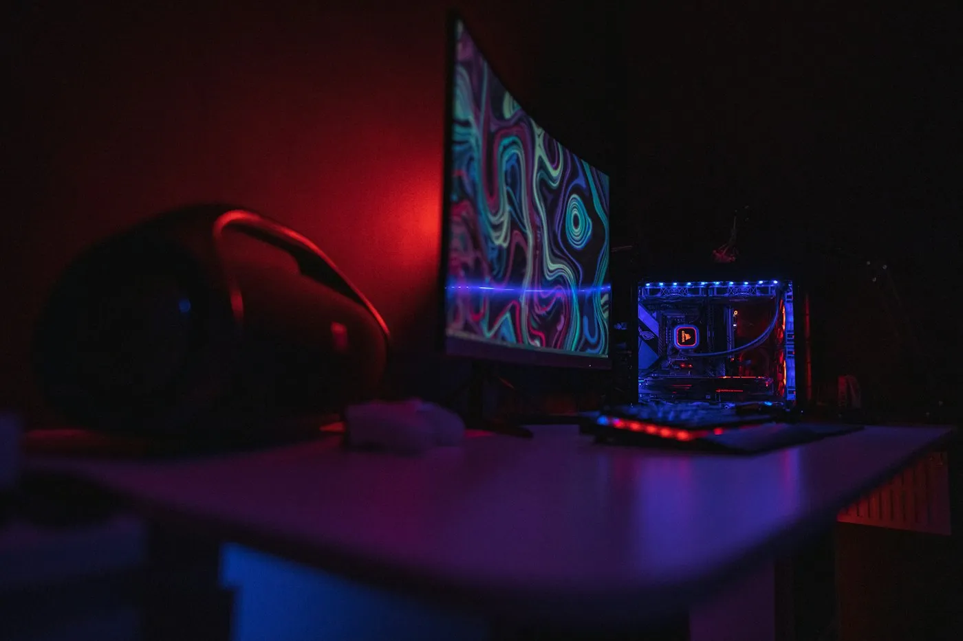 Dark gaming desk in red and blue light with a curved monitor, keyboard and glowing PC
