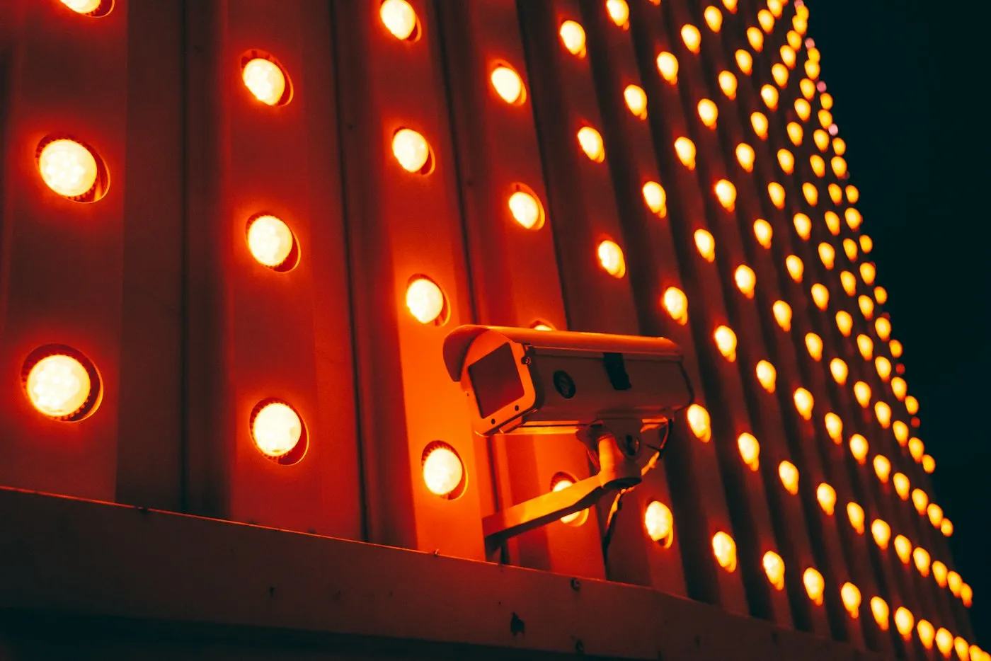 Bullet-style security camera mounted on a wall of glowing orange marquee lights at night